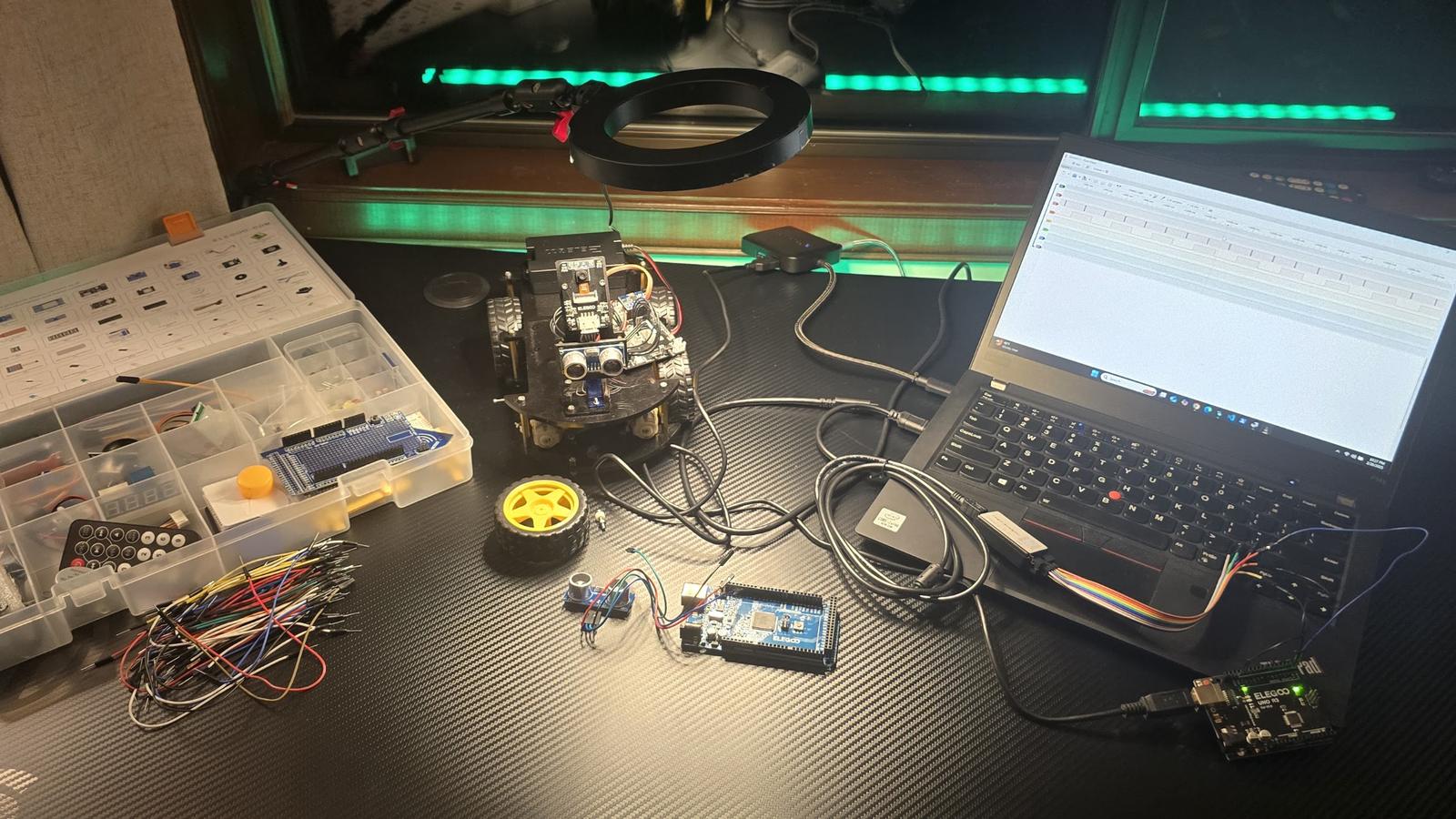 Workbench with an Arduino robot car, open sensor kit, jumper wires, and a laptop running the Arduino IDE